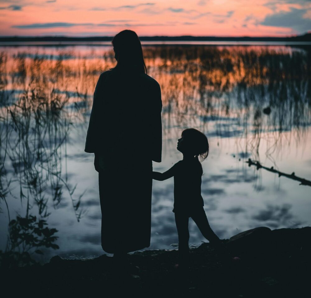 Woman holds hand with child in front of a body of water with a sunset.